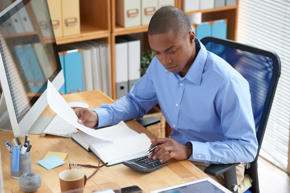 Fotografia de homem negro de cabeça raspada usando camisa social azul sentado em uma cadeira de escritório, calculando imposto de importação na calculadora enquanto segura uma folha de papel. Ele está em um escritório bem iluminado, com um monitor e teclado na mesa a frente dele.