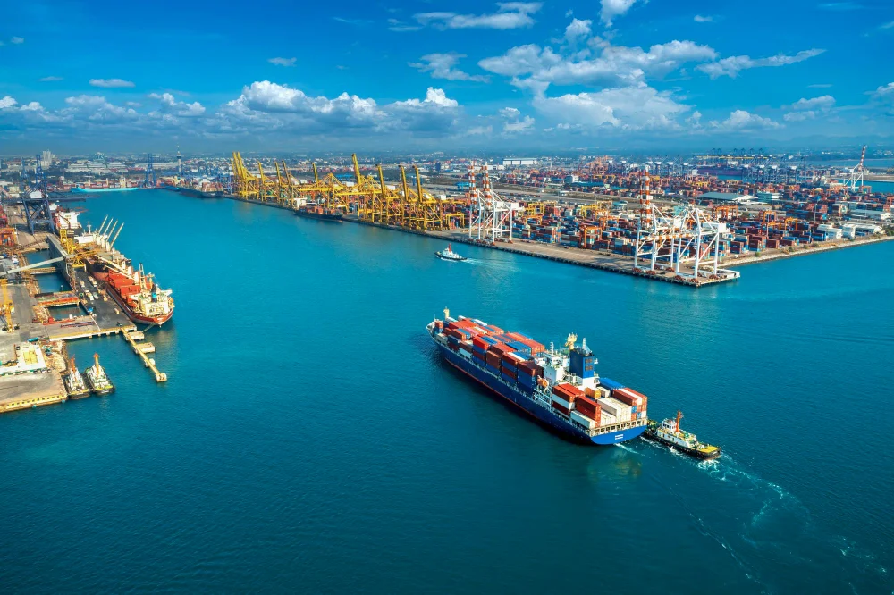 Fotografia de navio de carga entrando em porto, a fotografia é aérea e mostra parte do céu, que está azul com algumas nuvens